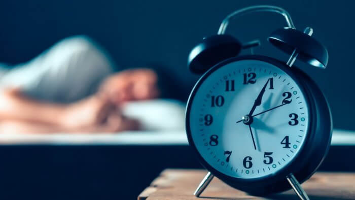 Alarm clock in the foreground with blurry photo of man asleep on bed behind