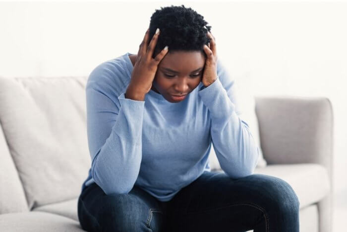 Worried looking lady of african origin sitting on sofa dressed in jeans and light blue blouse. - image depicts brain fog from long covid