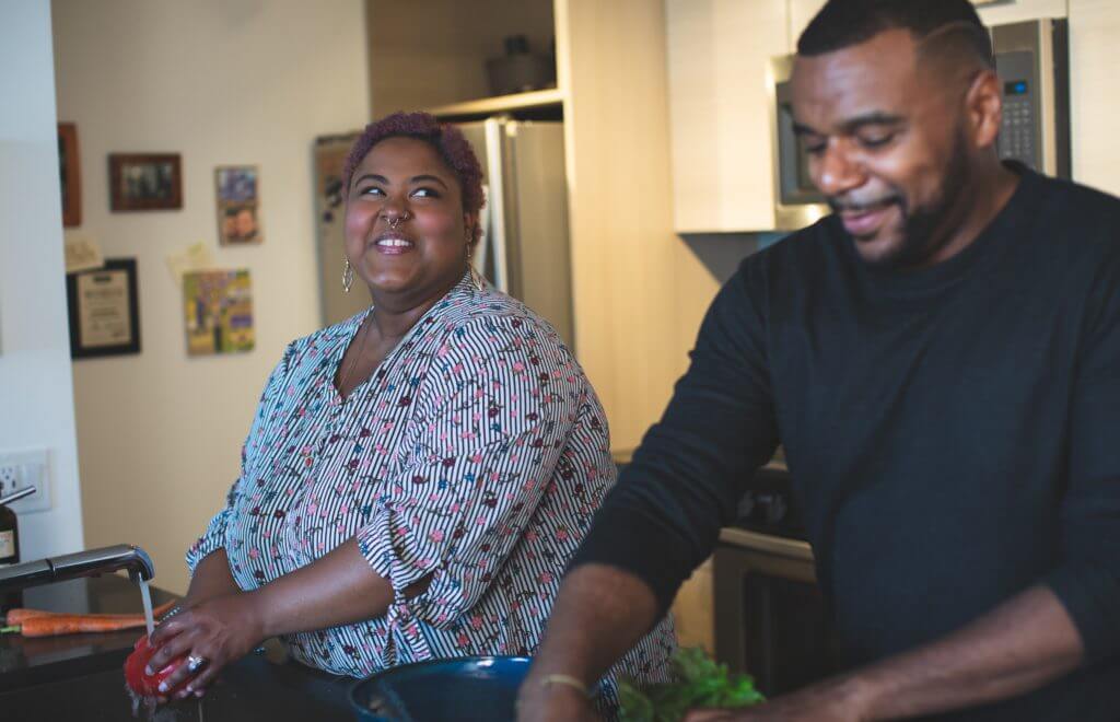 Overweight lady of African background standing at the sink and talking to her partner about safe contraception as they do the dishes.