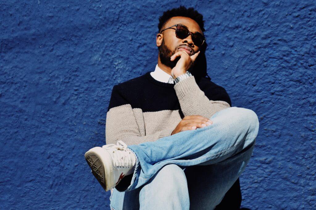 I snore - but why? Young man of African origin in sunglasses, jumper and jeans with his knee on the other leg.