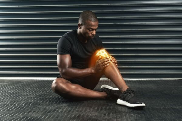 African man dressed in black sports gear holding to his knee which looks inflamed and painful
