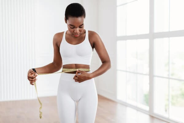 Cute black skinned lady in white workout gear and holding a yellow measuring tape around her weight - how well is her detox tea working?
