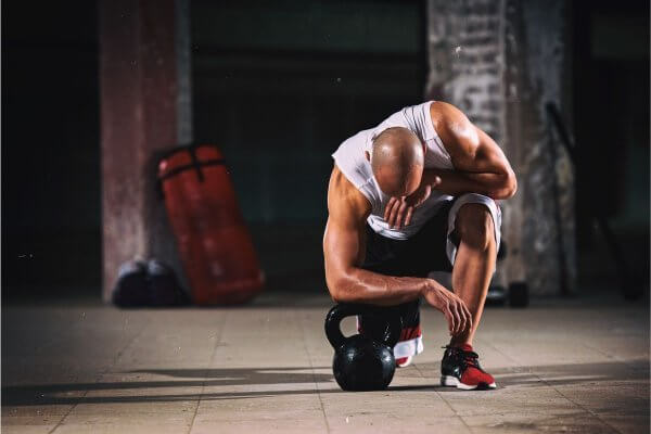Man of African origin on one knee in front of a kettle bell dressed in sleeveless top and shorts - appropriate dressing also key before you start your workout